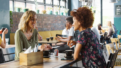 Two Young Female Friends Meeting Sitting At Table Drinking Hot Drinks Inside Coffee Shop And Talking
