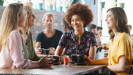 Multi-Cultural Group Of Female Friends Sitting At Table In Busy Coffee Shop Talking