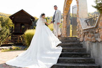 Obraz premium A bride and groom are standing on a set of stairs, with the bride wearing a white dress and the groom wearing a suit. The scene is set in a garden, with a wooden building in the background