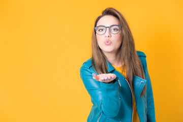 Lovely young woman send kiss to you, wearing blue leather jacket, isolated on yellow background....