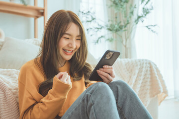 Excited young asian woman hold phone chatting in dating app feel happy sit on sofa at home, Closeup joyful female reading good news on phone. Surprised lady celebrating victory on phone.