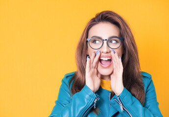Amused woman yelling, wearing blue leather jacket, isolated on yellow background. Happy girl holding hands near his mouth
