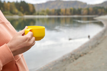 Relaxing morning by the lake: close up of hand holding yellow mug with tranquil waters and scenic forest background.