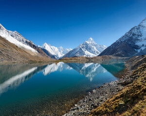 Calm lake at the base of snow-capped mountains, mirroring the clear blue sky on a sunny day, ai.