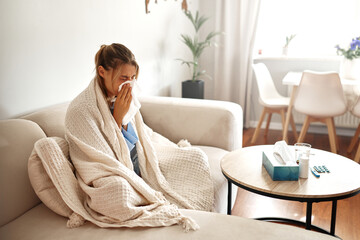 A young woman feeling unwell is wrapped snugly in a blanket while sitting on her couch