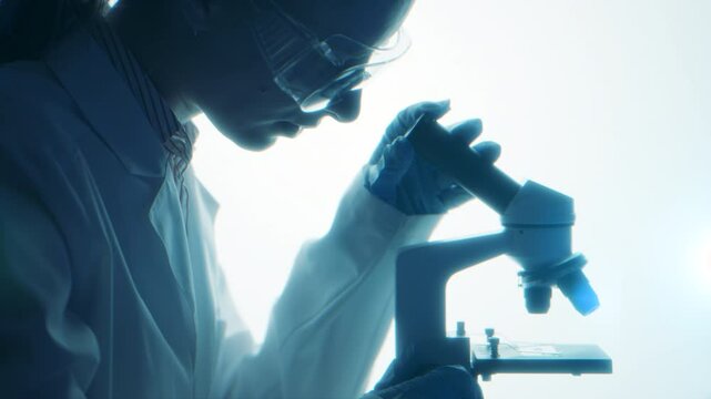 Blue toned silhouette of a scientist bent over the microscope. Cold laboratory light and quiet work