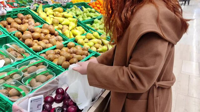 Redhead Woman Purchasing Organic Kiwi Fruits at a Supermarket Store, Grocery Department. Young Lady Picks Fruits and Put them in a Bag. Fruit Display in Crates at the Local Market. 