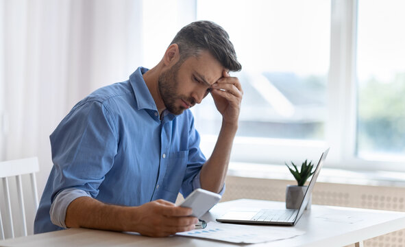 Worried male freelancer looking at smartphone screen while working at home office, received bad news, got scam message, thoughtful man touching forehead with concerned face expression, copy space