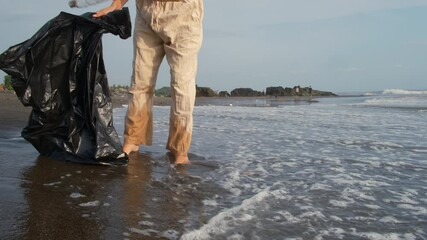 A woman cleans up a beach by collecting litter in a large black bag. The serene sea and gentle waves contrast with the environmental pollution. A volunteer collects plastic waste and other litter.