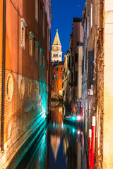 A narrow canal in Venice, Italy, at night, with a gondola passing under a bridge and a tall bell tower of St Mark's Campanile visible in the distance.