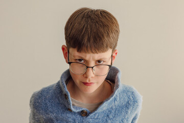 angry teenager, young boy frowning over his glasses child, 11 year old , wearing knitted wooly blue jumper, brown hair, caucsian, brown eyes, serious face