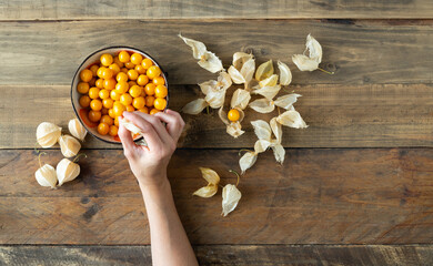 a hand taking a golden berry from a bowl. Around there are fruits and leaves. Physalis peruviana