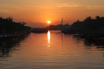 Sunrise over a calm river in Hoi An, Vietnam, with the sun casting a warm reflection on the water and silhouettes of trees and boats.