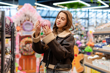 Fototapeta premium A young woman with long hair, wearing a short brown sheepskin coat, is choosing Christmas ornaments in a store. She is holding a pink Christmas ornament in her hands.