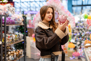 A young woman with long hair, wearing a short brown sheepskin coat, is choosing Christmas ornaments in a store. She is holding a pink Christmas ornament in her hands.