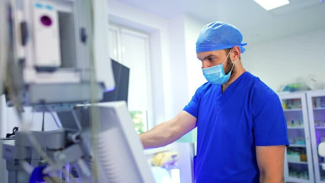Bearded medic in blue uniform stands at the medical equipment. Doctor pressing the keys on the machine during operation.