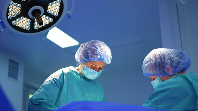 Two female medics in masks, caps and blue robes under the round lamp. Low angle view at the doctors at surgery.