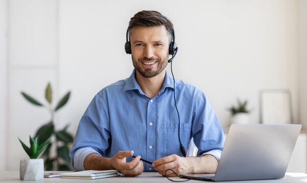 Portrait of smiling male customer service operator in headset posing at workplace, looking at camera, portrait of handsome hotline manager sitting at desk with laptop in white office, copy space