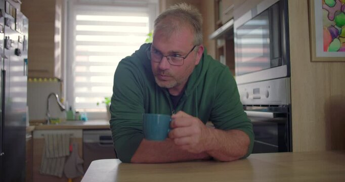 Man seated at kitchen counter with mug, reflective pose, domestic lifestyle, modern home environment, casual morning routine with natural light and greenery