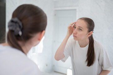 Obraz premium Unhappy gen z woman finding an acne on forehead, stands before a mirror in the bathroom. Concept of acne treatment, skincare, adult acne