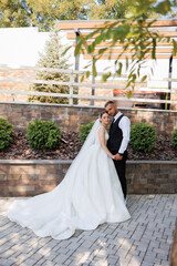 A bride and groom are posing for a picture in front of a brick wall. The bride is wearing a white dress and the groom is wearing a black vest. Scene is happy and celebratory