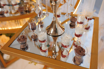A table with a variety of desserts, including cakes and cupcakes, is displayed on a gold tray. The desserts are arranged in small cups and are surrounded by a mirror, creating an elegant