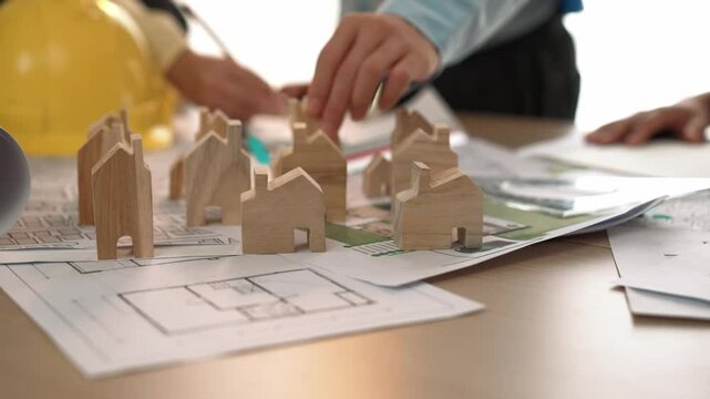 Architect and engineer discuss about building blueprint on table with architectural document, measurement tools scatter around at office, use wooden block for house positioning. Closeup. Delineation.
