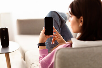 Over The Shoulder View Of Unrecognizable Young Woman Holding And Using Cell Phone With Empty Mockup Screen, Sitting On Couch In Living Room At Home. Selective Focus On Gadget, Template