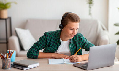 Joyful teen guy writing at notebook and looking at laptop screen, having lesson online, home...