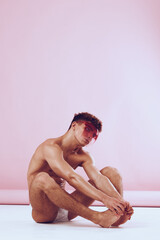 Obraz premium Thoughtful young man sitting on the floor, surrounded by a soft pink background, expressing calm and introspection. Fashion and emotional expression concept.