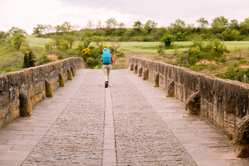 A traveler walks alone across a historic stone bridge in Obanos, Spain, surrounded by nature