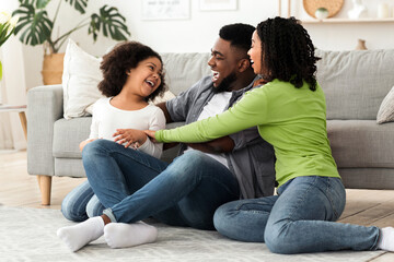 Cheerful black family having fun at home, parents tickling their daughter, laughing together,...