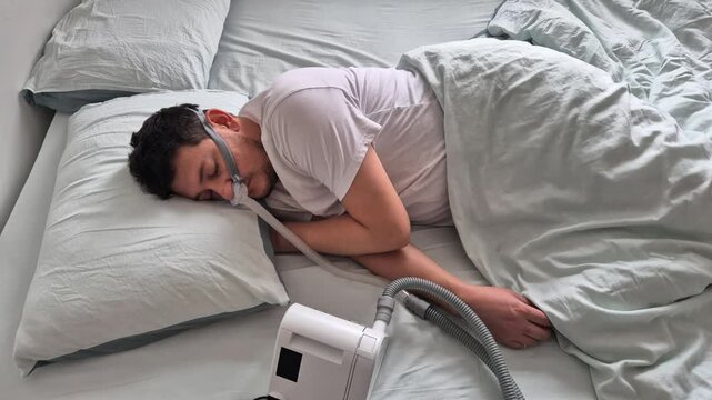 A man in his mid-thirties with sleep apnea disorder, sleeps using a nasal mask and CPAP device in a bed in his house