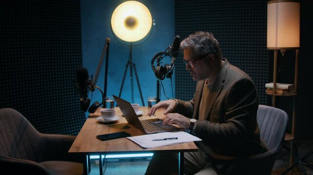 Medium shot of middle-aged Caucasian male business podcast host sitting at table in studio with microphones and headphones, getting ready for show, researching interview questions online on laptop