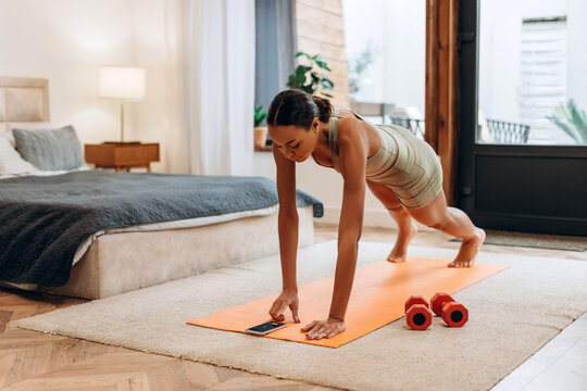 Fit African American woman doing plank exercise, turning on the timer on mobile phone at home
