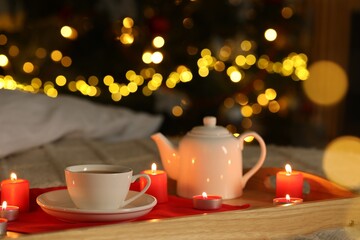 Aromatic tea in cup, teapot and burning candles on bed against blurred Christmas lights, closeup. Bokeh effect