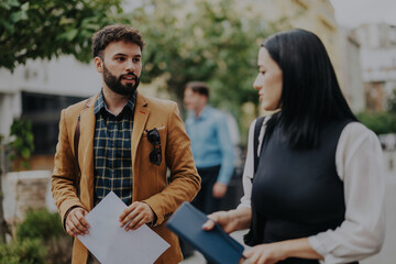 Two young business people engage in a lively conversation outdoors in the city. The urban backdrop indicates a bustling atmosphere, perfect for networking and collaboration.