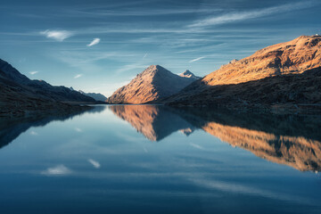 Sunset casts a serene glow on mountain peaks mirrored in a tranquil alpine lake in autumn. Lake Bianco, Switzerland in fall. Landscape with mountains, reflection in water, blue sky. Picturesque scene