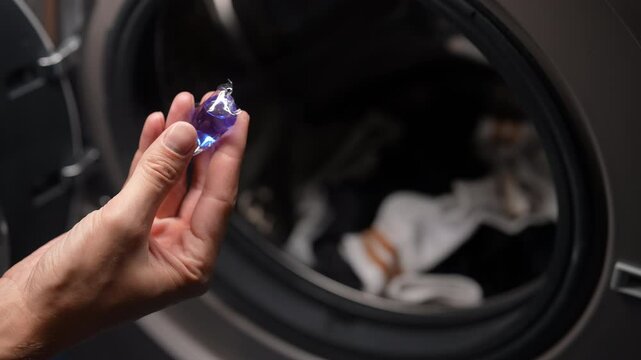 Male hand holding and throwing blue liquid capsule with laundry detergent into washing machine tank filled with clothes, ready to start wash cycle. Concept of easy housekeeping domestic life.