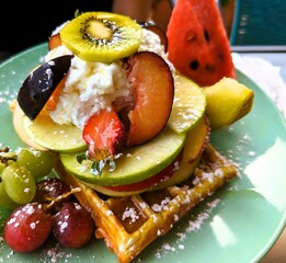 Belgian waffle with fruit salad garnished with icing sugar and whipped cream, sweet breakfast and desert, close-up