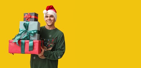 Handsome young man in Santa hat holding Christmas gifts on yellow background with space for text