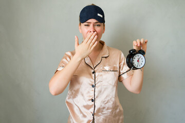 Sleepy woman in pajamas and eye mask yawning while holding an alarm clock
