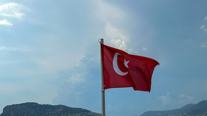 A Turkish flag waving against a blue sky with soft clouds, set against a backdrop of a scenic mountainous landscape. Highlights national pride, patriotism, and Turkish culture.