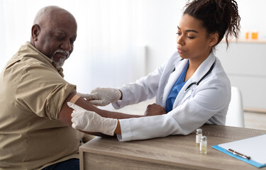 Vaccination Concept. Senior Black Man Getting Vaccinated Against Flu Or Corona Virus With Antiviral Vaccine, Female Doctor Putting Applying Sticking Adhesive Bandage On Arm After Injection In Hospital