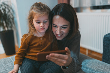 A joyful mother and her adorable daughter spend quality time indoors, exploring content on a smartphone. The image depicts a warm and loving family moment.