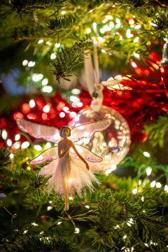 Christmas tree fairy with gossamer wings balances on a branch with crystal bauble and fairy lights in the background. Festive scene.
