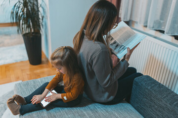 In a tranquil home setting, a mother reads a book while her child is occupied with a smartphone, each engaged in their own world of exploration and learning.