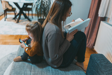 A woman reads a book while a young child is engrossed in a smartphone, sitting back-to-back on a...