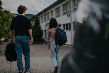 A parent drops off their daughter and son at school. The children are walking towards the building, carrying backpacks. It captures the start of a school day routine.