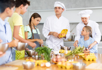 Amiable woman and young man, skilled chefs wearing white cook jackets and toques, running culinary courses for preteen children, sharing secrets of cooking